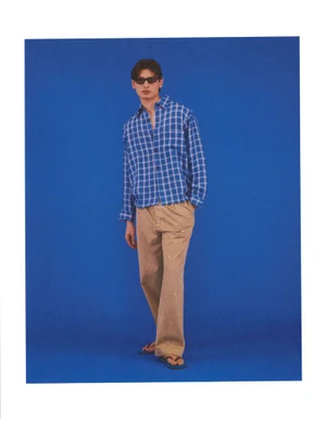 Model wearing dark sunglasses, a blue plaid button-down shirt, and beige trousers against a vibrant blue studio backdrop.