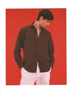 Model wearing a brown button-up shirt and white trousers against a vibrant red backdrop.