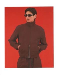 Ellis Mueller wearing a dark brown zip jacket and black sunglasses with hands in pockets against a bright red studio background.