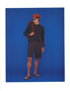 Model Ellis Mueller wears a navy long-sleeve shirt, dark shorts, sandals, and a red cap against a bold blue studio backdrop.