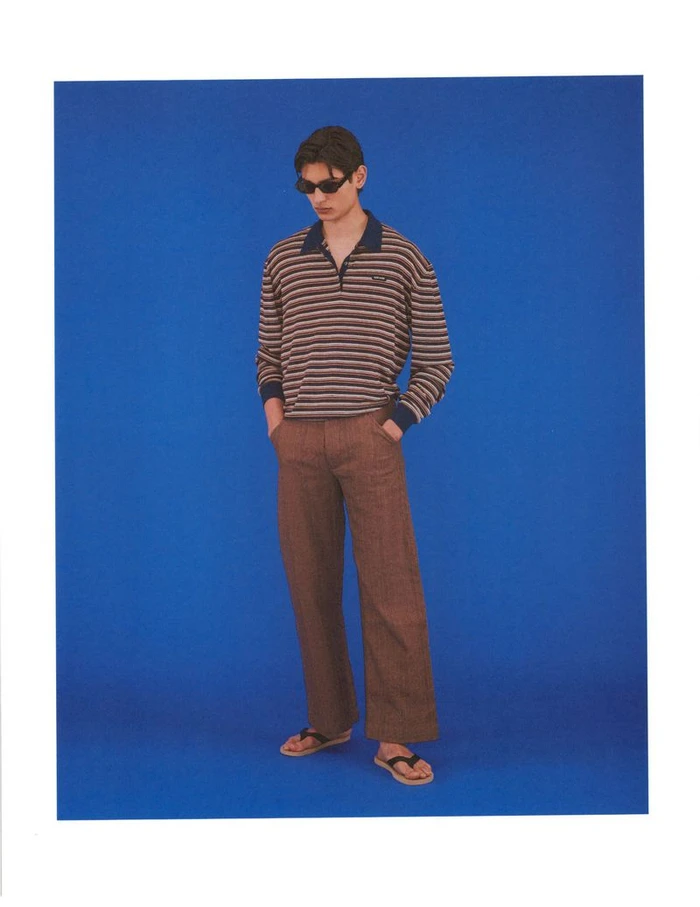 Model Ellis Mueller standing in sunglasses, a striped collared long-sleeve shirt, and brown trousers against a solid blue backdrop.