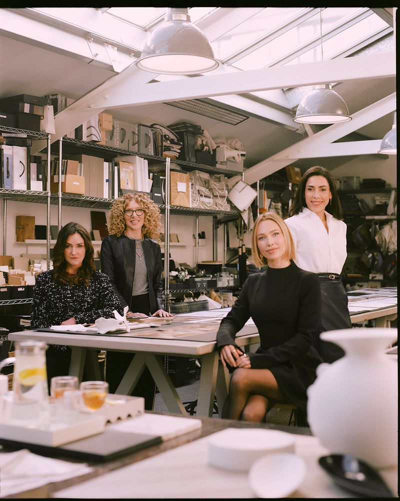 Four women posing in a design studio workshop with shelves of material samples and architectural models in the background.