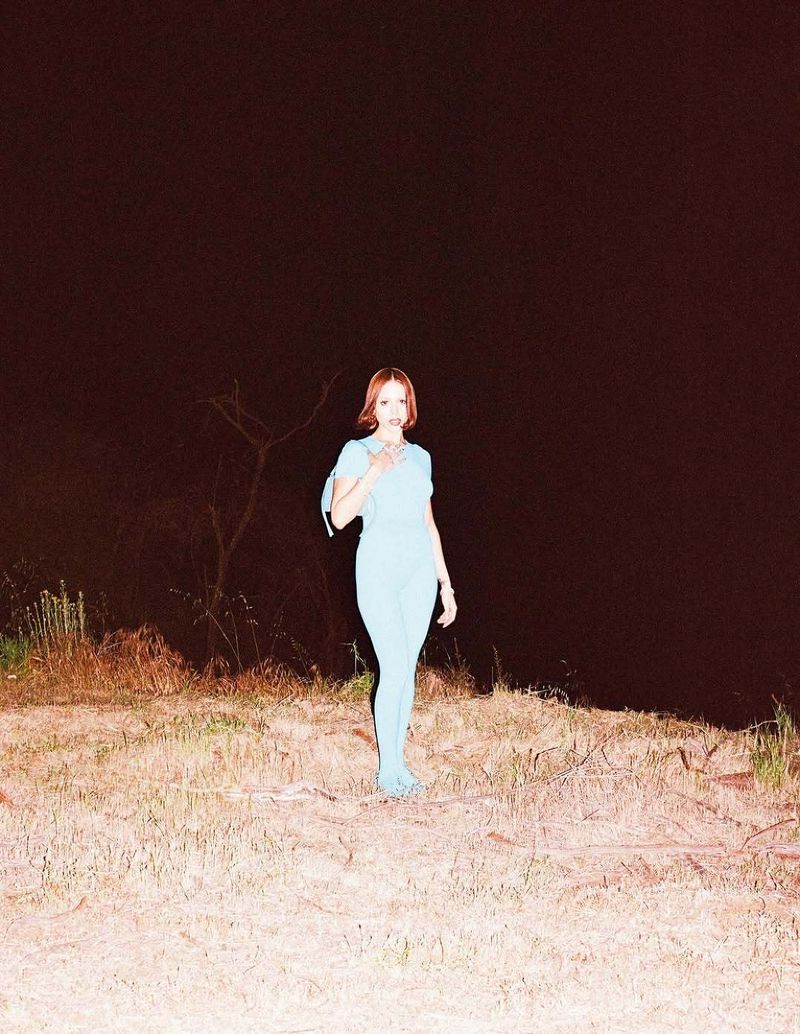 Woman standing in a dry grassy field at night illuminated by flash, wearing a powder blue matching outfit and shoulder bag.