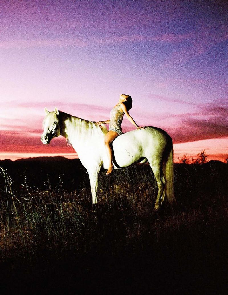 Woman in a shimmering mini dress leaning back on a white horse in a field under a vivid purple and pink sunset sky.
