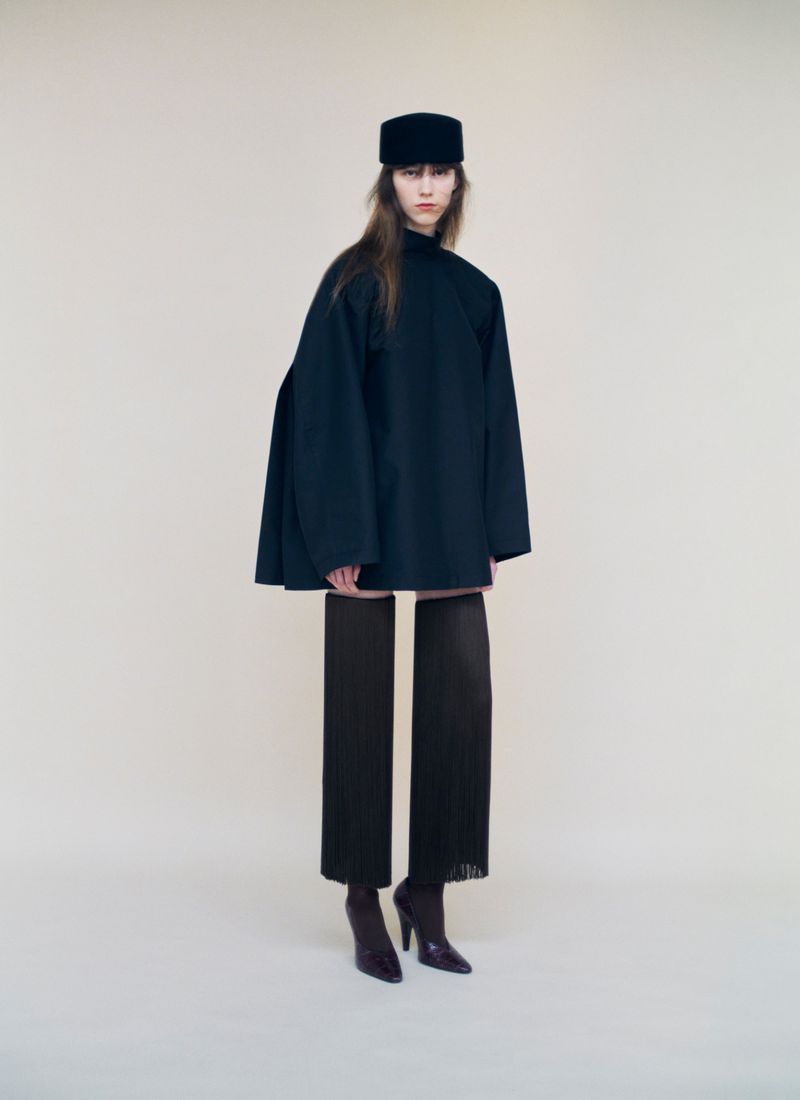 Model Kaat Van Herbruggen posing in an oversized black structured cape coat, black pillbox hat, and fringe trousers on a pale background.