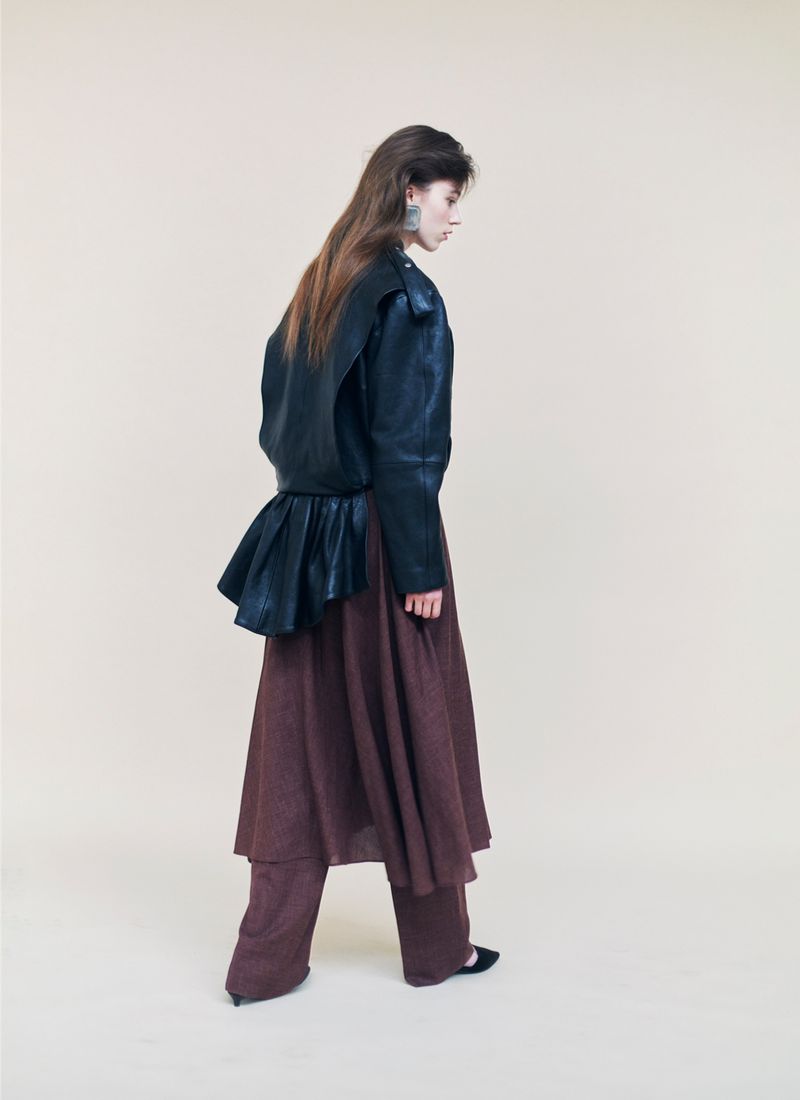 Model Kaat Van Herbruggen wearing a black leather jacket and brown trousers against a seamless neutral studio backdrop.