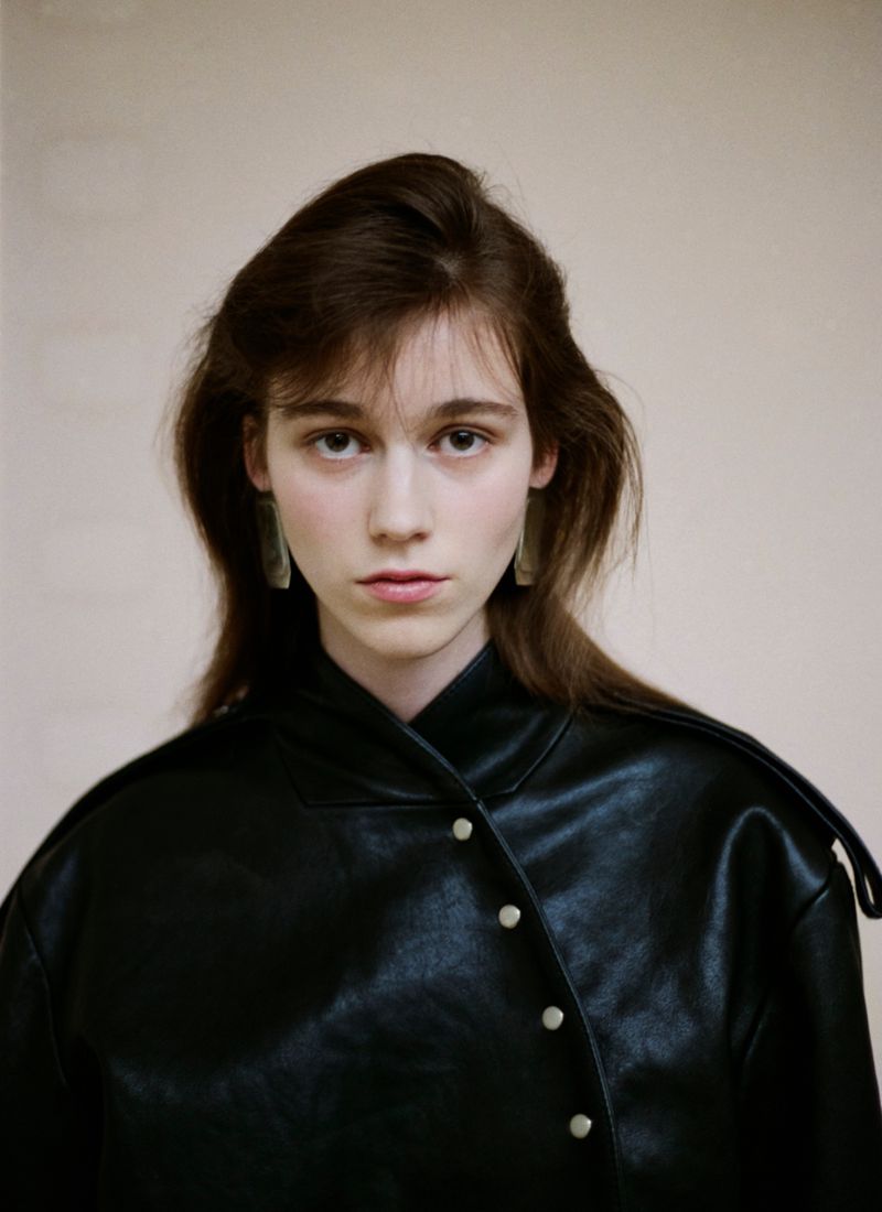 Portrait of a woman wearing a black leather buttoned jacket with long brown hair against a neutral backdrop
