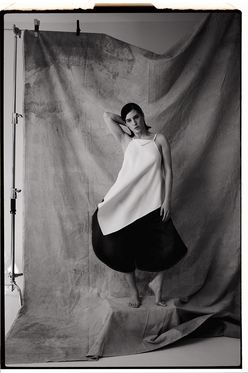 Monochrome fashion portrait of a woman posing barefoot in an asymmetrical white top and voluminous black skirt before a draped backdrop.