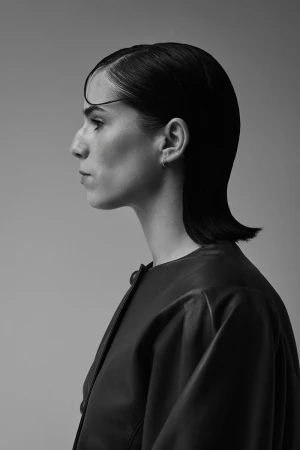 Monochrome profile portrait of a woman with slicked-back dark hair wearing a black leather jacket against a neutral studio backdrop.