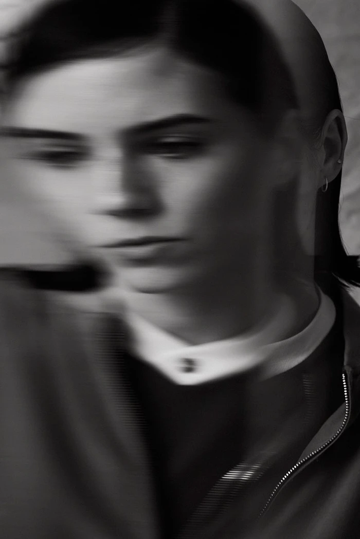 Black and white motion-blurred layered portrait of a woman wearing a dark zip-up jacket and white collared shirt.