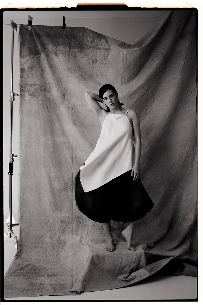 Monochrome fashion portrait of a woman posing barefoot in an asymmetrical white top and voluminous black skirt before a draped backdrop.