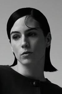 Black and white close-up portrait of a woman with slicked-back dark hair looking to the side against a neutral studio background.