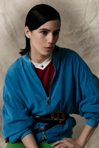 Model with slicked hair wearing a bright blue bomber jacket and layered black belts against a neutral textured studio backdrop.