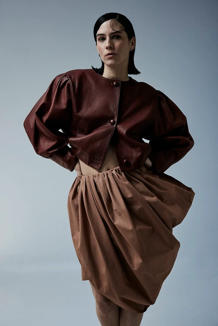 Model posing in a cropped burgundy puff-sleeve leather jacket and voluminous tan draped skirt against a cool grey studio backdrop.