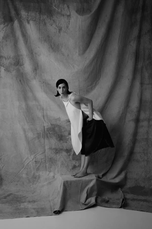 Monochrome studio portrait of a woman in an asymmetrical white top and dark culottes standing barefoot on a draped pedestal.