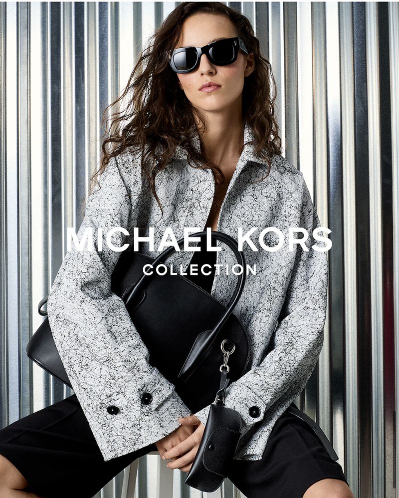 Model Emm Arruda wearing sunglasses and patterned jacket holding black handbag for Michael Kors Fall/Winter 2026.