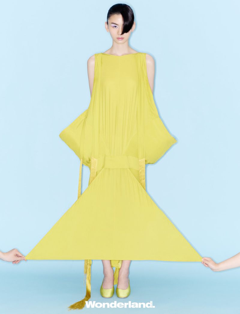 Model posing in an architectural yellow dress held taut into a geometric silhouette against a pale blue backdrop.