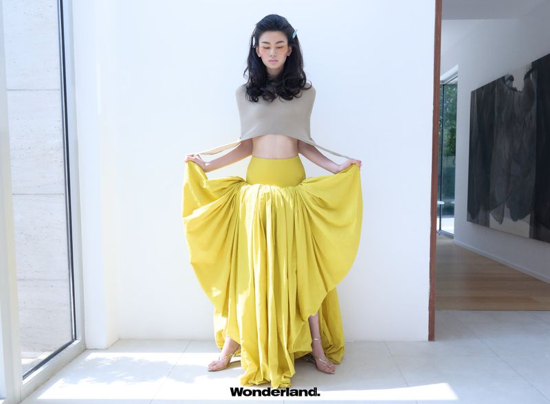 Model wearing a beige cropped top and holding a voluminous draped yellow maxi skirt in a bright hallway.