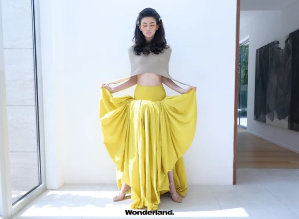 Model wearing a beige cropped top and holding a voluminous draped yellow maxi skirt in a bright hallway.
