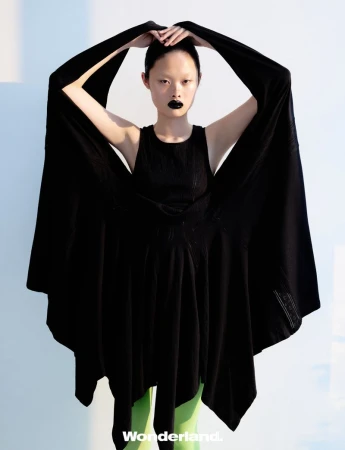 East Asian model in a voluminous black draped dress and bright lime green tights posing with raised arms against a pale backdrop.