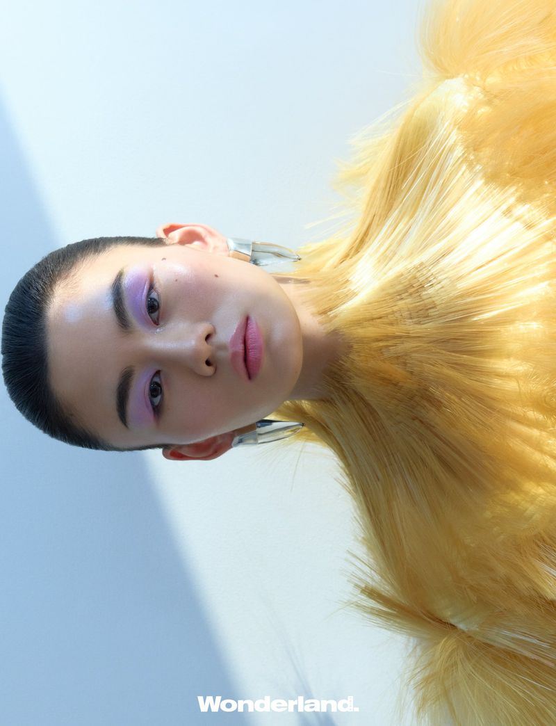 Low-angle beauty portrait of an East Asian model with lilac eye makeup wearing a yellow textured garment and silver earrings.