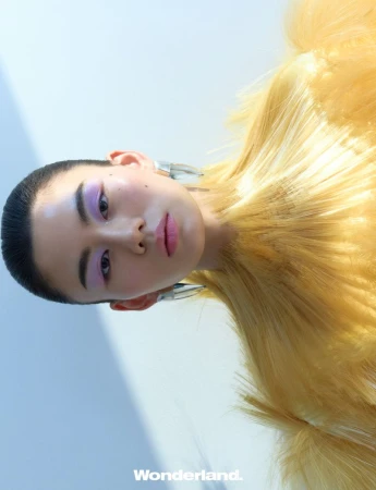 Low-angle beauty portrait of an East Asian model with lilac eye makeup wearing a yellow textured garment and silver earrings.