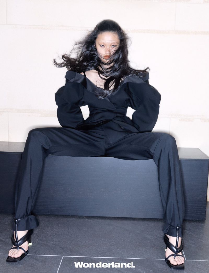 Model seated wearing a black off-the-shoulder structured suit with black heeled sandals against an indoor wall.