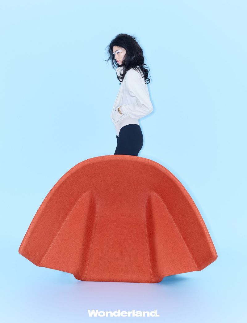 Model in a white jacket standing behind a large sculptural orange chair against a light blue studio backdrop.