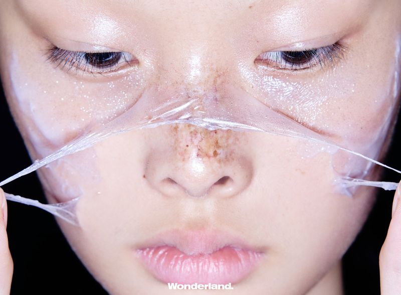 Close-up of an East Asian model peeling a translucent face mask across her nose and cheeks against a dark studio background.