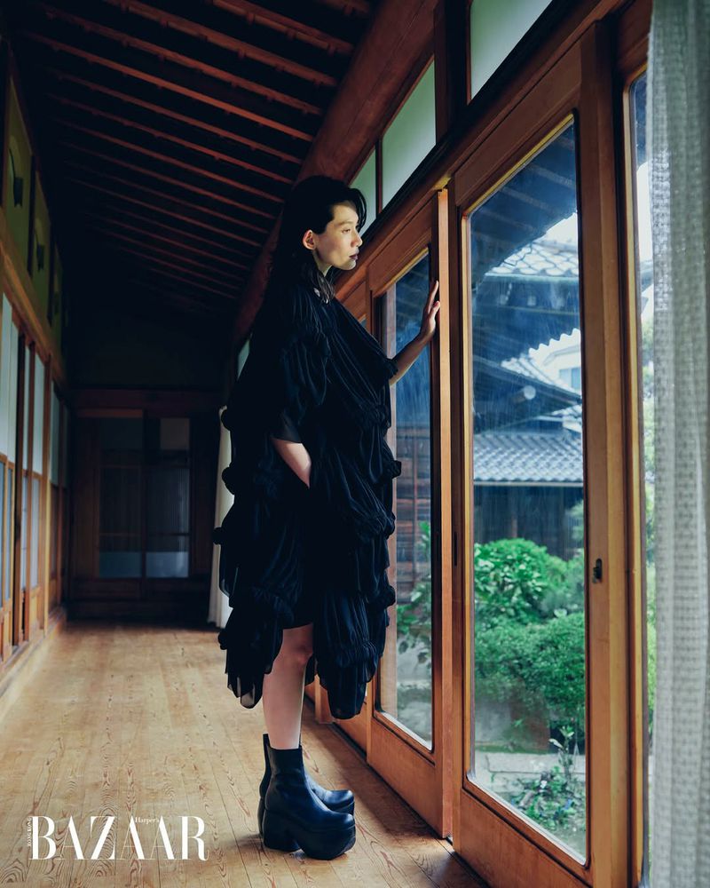 Woman in an oversized tiered black dress and chunky platform boots standing by glass sliding doors in a wooden corridor.