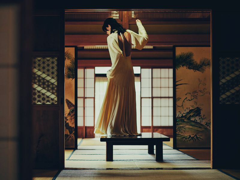 Model standing on a low dark wooden table in an off-white backless gown inside a traditional Japanese tatami room with shoji screens.