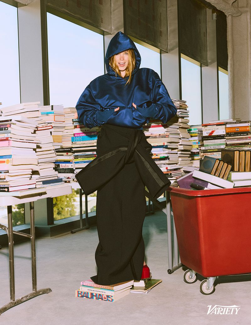 Milly Alcock wearing a navy hooded jacket and black draped skirt standing on a stack of books for Variety Magazine.