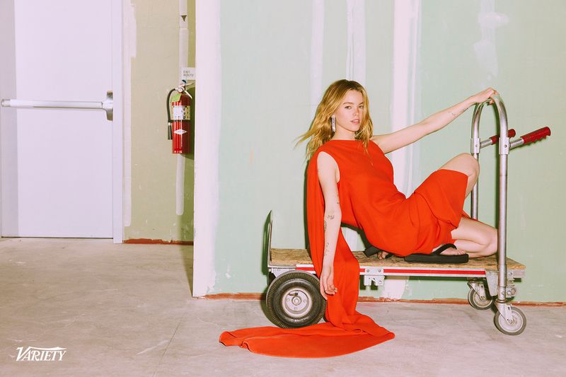 Milly Alcock wearing a long red draped dress and black platform sandals while posing on a metal utility cart against green drywall.