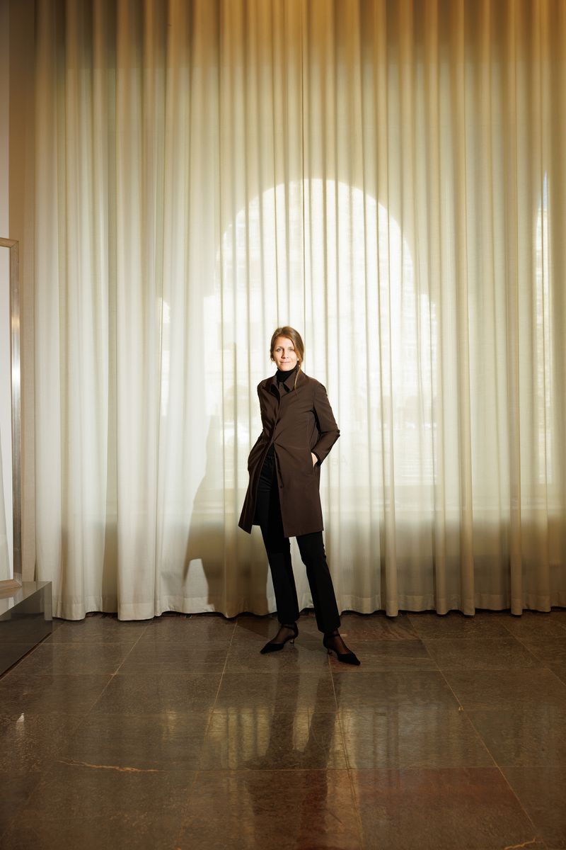 Luisa Dames standing in a tailored dark coat and black trousers in front of floor-to-ceiling pleated cream curtains.