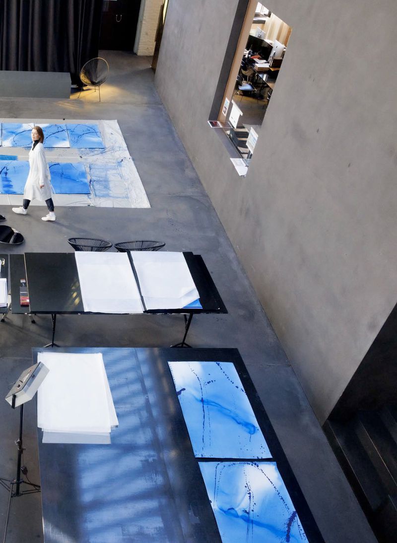 High-angle view of artist Jorinde Voigt in a white coat walking across a spacious studio filled with blue artwork tables.