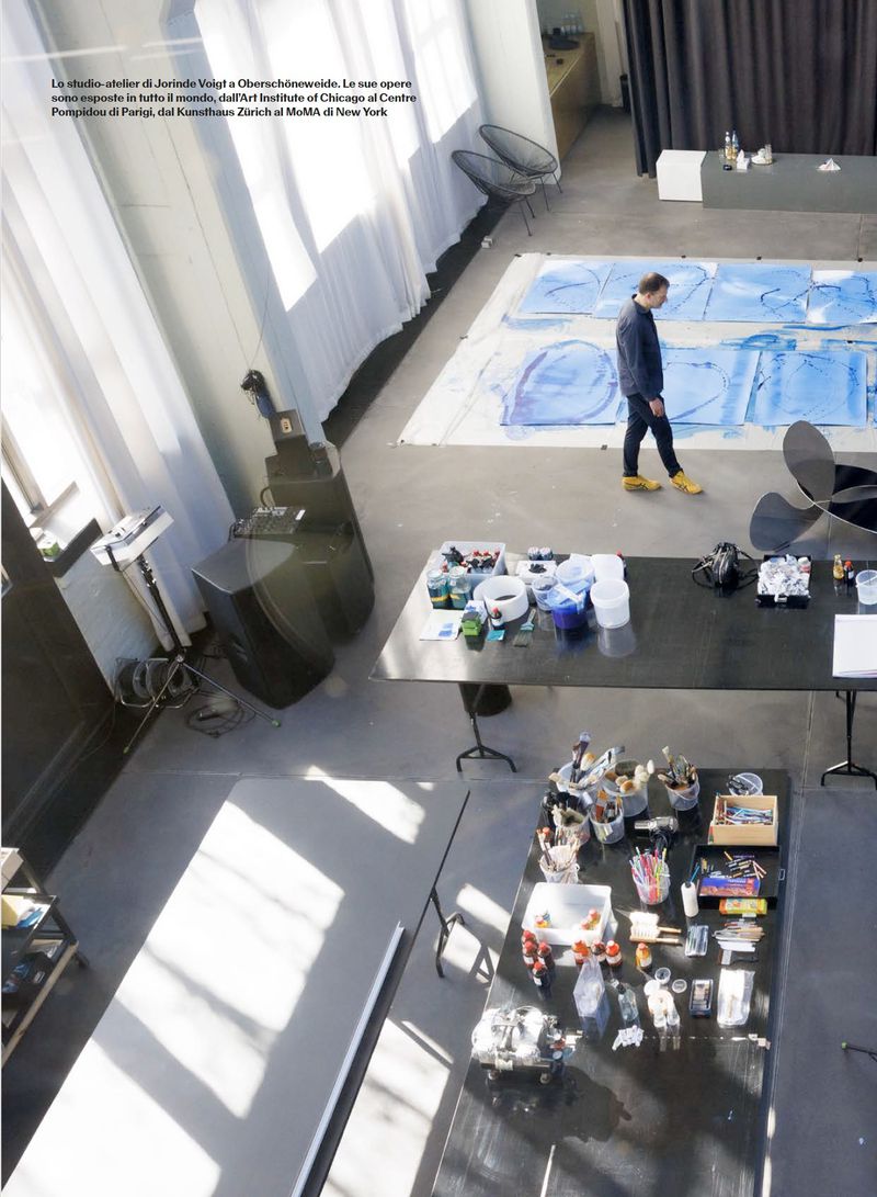 High-angle editorial view of an artist walking through a bright studio workspace next to large blue artworks on the floor