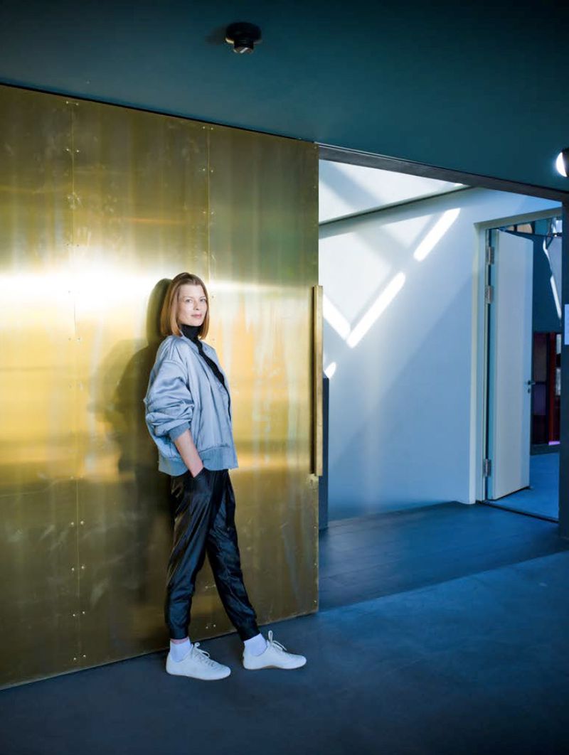 Jorinde Voigt leans against a large gold brass sliding door wearing a grey bomber jacket, black trousers, and white sneakers.