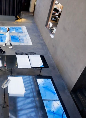 High-angle view of artist Jorinde Voigt in a white coat walking across a spacious studio filled with blue artwork tables.