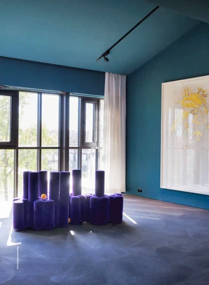 Sculptural purple cylindrical sofa in a modern teal room with large windows, white sheer curtains, and framed yellow artwork.
