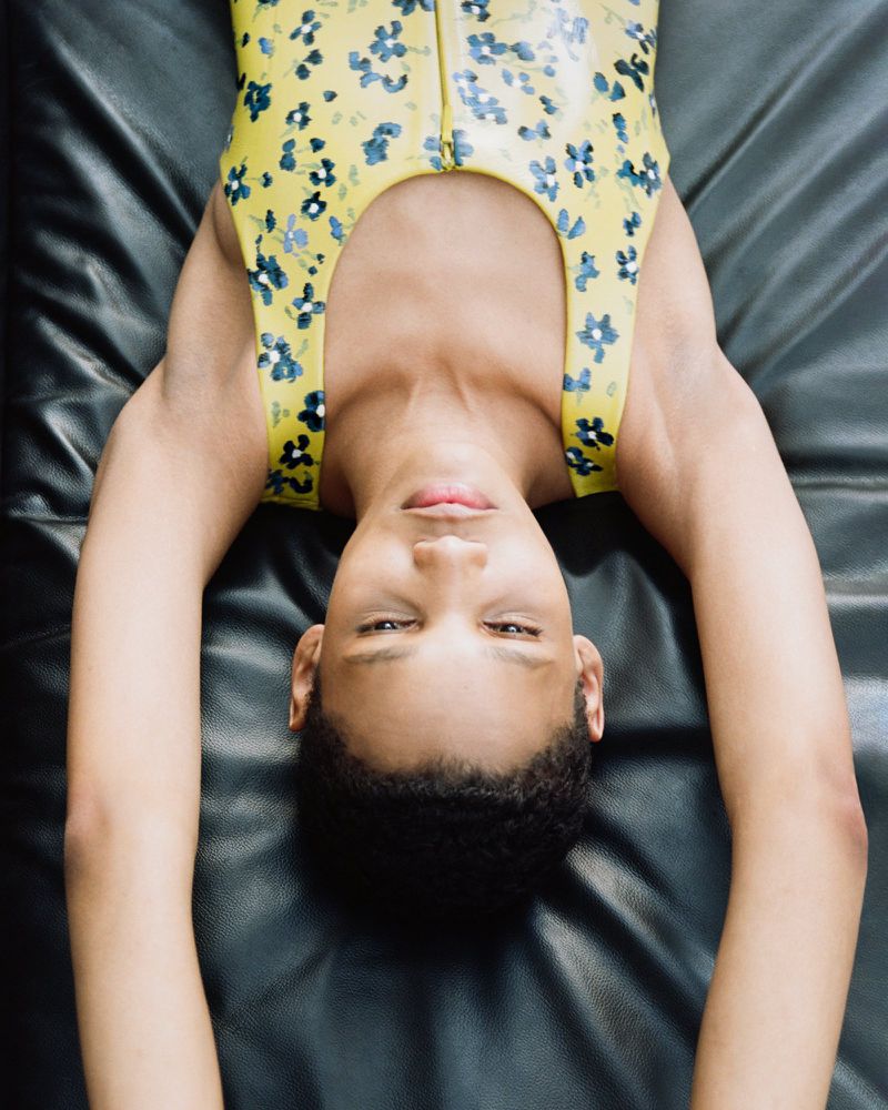Model Haojie Qi lying upside down on black leather wearing a yellow floral sleeveless bodysuit.