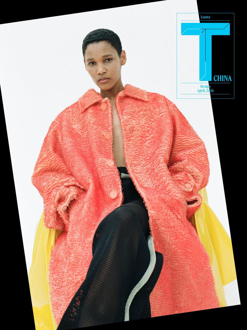 Model seated wearing an oversized textured coral coat and black mesh trousers against a white seamless backdrop.