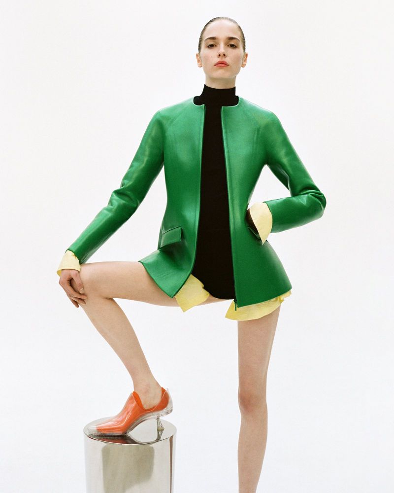 Model wearing a structured green jacket and black high-neck top posing with one foot on a chrome plinth against a white background