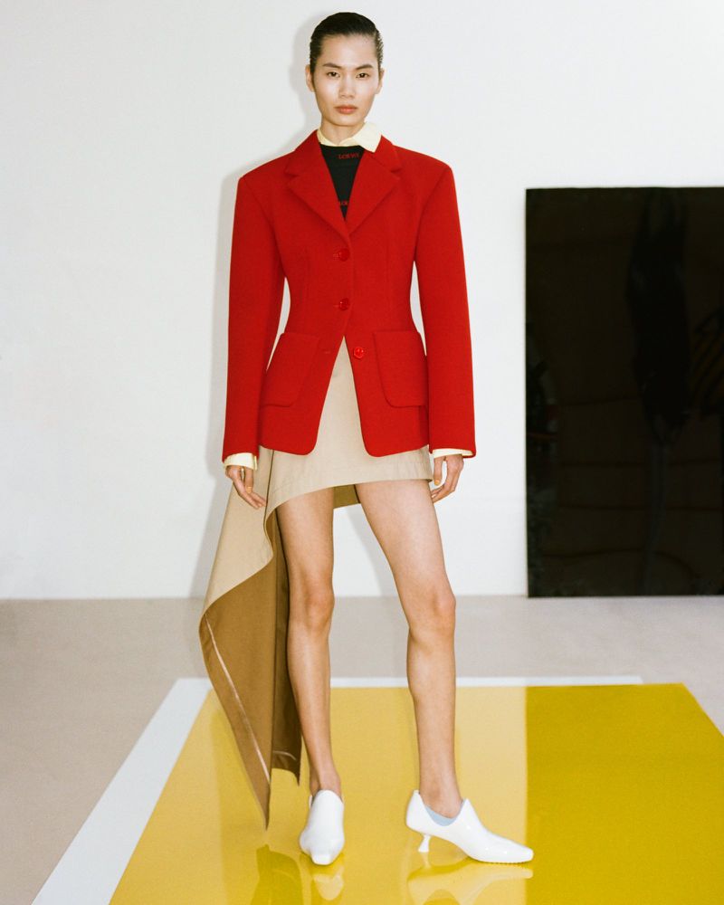 Model wearing a red structured blazer and asymmetric beige skirt standing on a reflective yellow platform