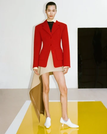 Model wearing a red structured blazer and asymmetric beige skirt standing on a reflective yellow platform