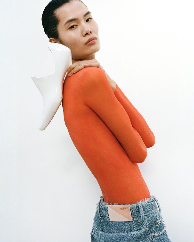 Model Haojie Qi wearing an orange long-sleeve top and frayed denim while resting a sculptural white heel on her shoulder.