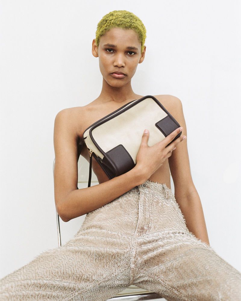 Model with short chartreuse hair seated holding a cream and dark brown handbag wearing textured beige pants against a white background.