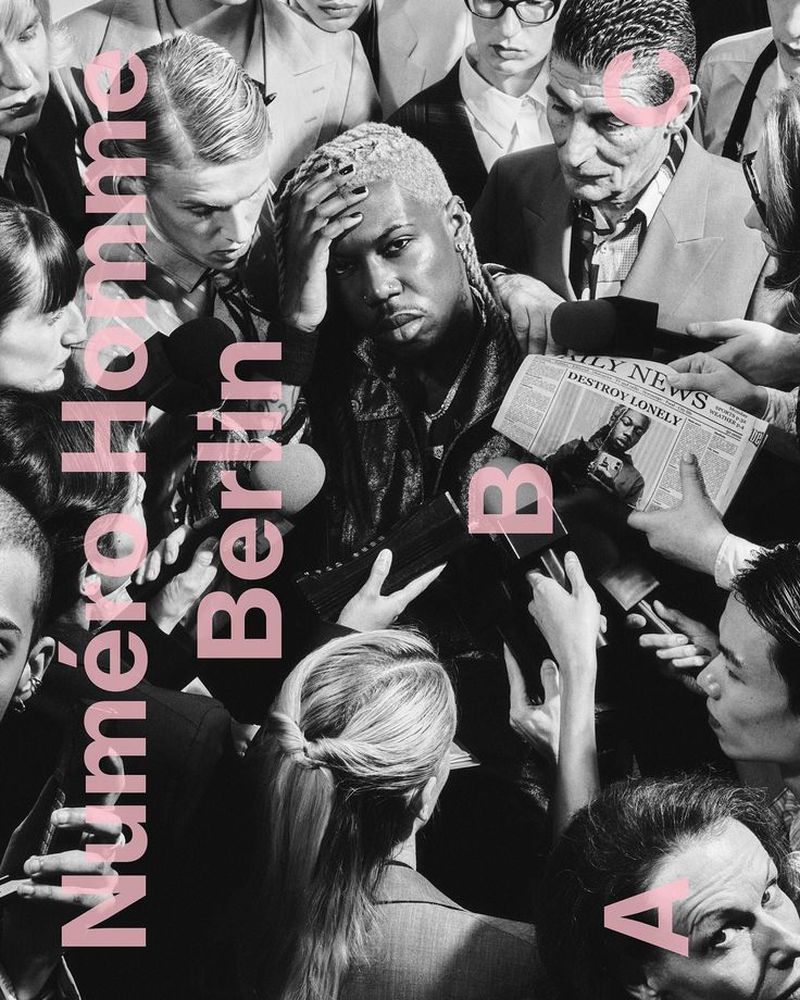 Editorial fashion shot of a man surrounded by people holding microphones and newspapers, with pink text overlay on a monochrome image.