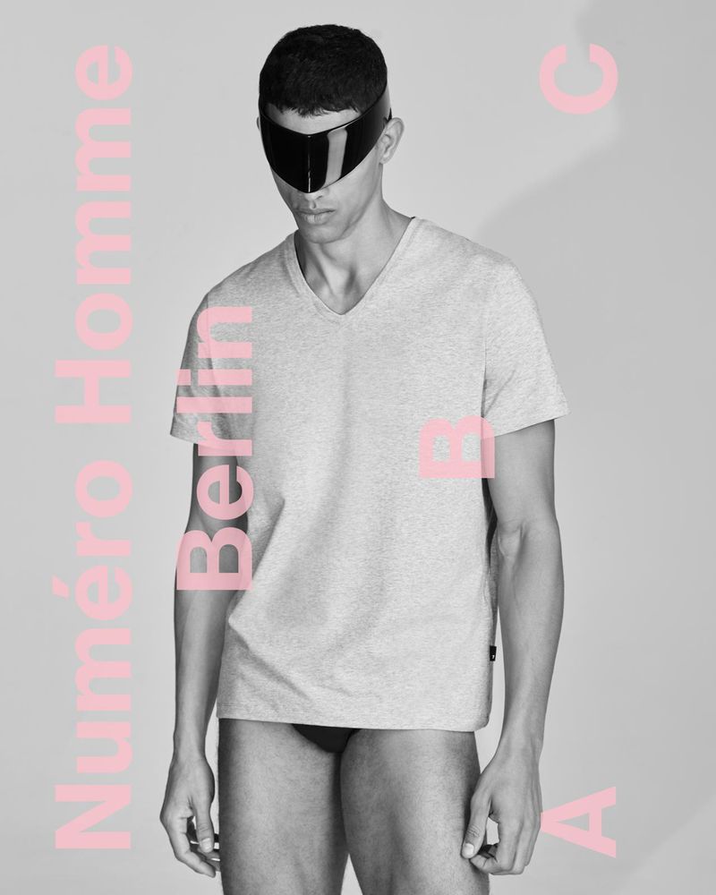 A man in a grey t-shirt poses indoors with pink text overlay. The image showcases a studio shot emphasizing fashion and minimalist aesthetics.