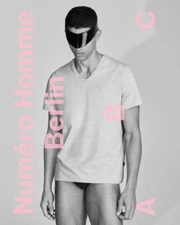 A man in a grey t-shirt poses indoors with pink text overlay. The image showcases a studio shot emphasizing fashion and minimalist aesthetics.