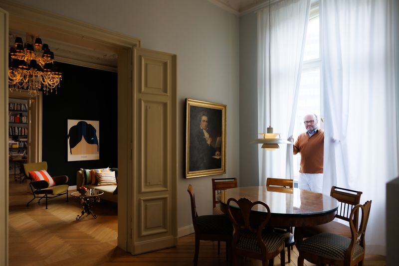 Thomas Demand standing by sheer white curtains in a stylish dining room featuring a round wooden table and vintage chairs.